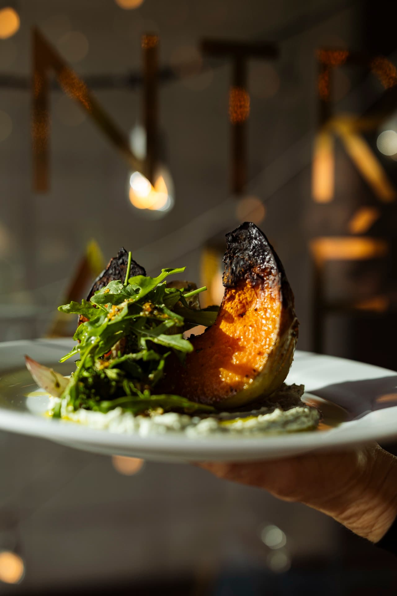 A waiter holding a plate of charred ember-roasted pumpkin under the brass MANTRA letters